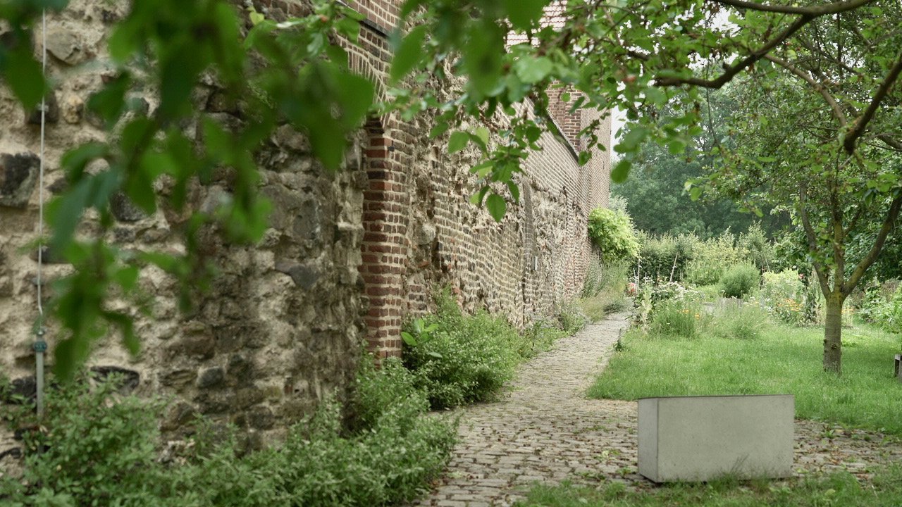 Ein gepflasterter Pfad entlang einer alten, grün bewachsenen Mauer, auf der anderen Seite eine Wiese mit Bäumen.