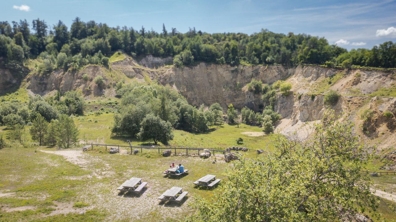 Sicht von einer Anhöhe auf eine Ebene die von dicht bewachsenen Felsen umgeben ist. Unten stehen Picknickbänke.