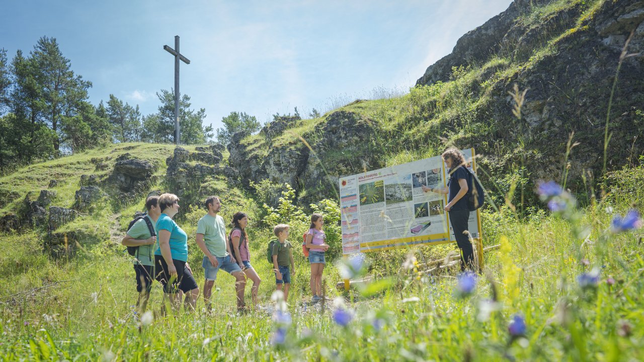 Eine Besuchergruppe steht vor einer Informationstafel auf einer grünen, blumigen Wiese. Im Hintergrund sind grün bewachsene Felsen zu sehen, oben steht ein Holzkreuz.