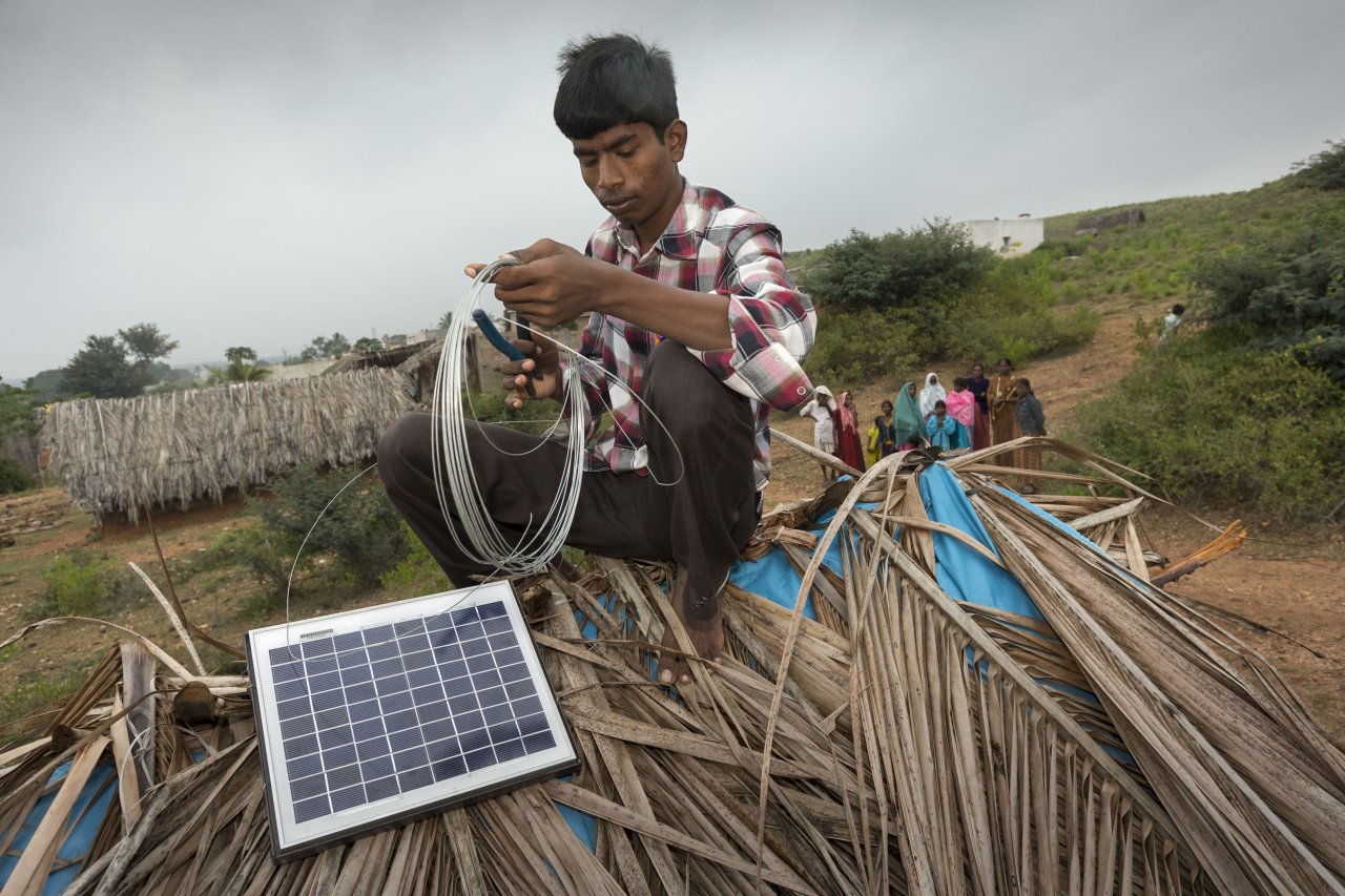  Auf dem mit Palmwedeln gedeckten Dach der Hütte von Frau Sharamamma positioniert der Mechaniker Shivakumar das Solarpanel. Im Hintergrund beobachten Dorfbewohner das Geschehen.