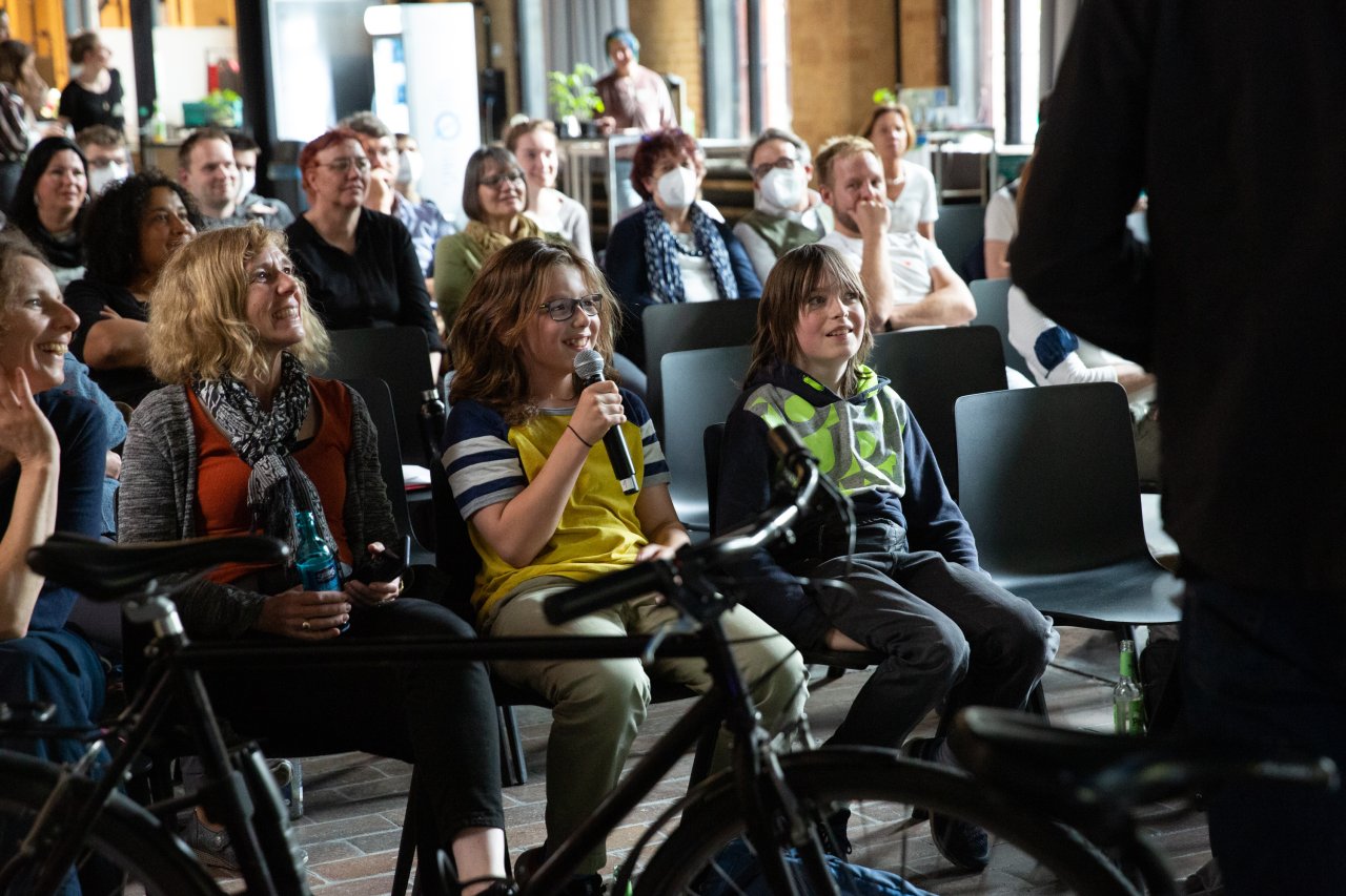 Blick ins Publikum: Teilnehmende schauen gebannt nach vorne, während ein Kind aus dem Publikum das Mikro hält und spricht.