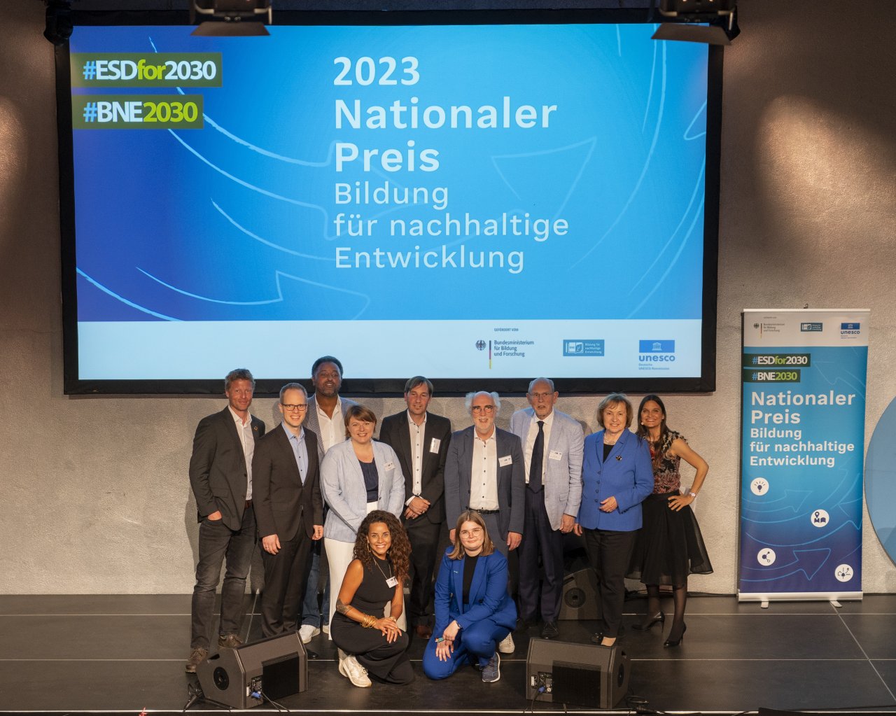 Mitglieder der Jury des BNE-Preises, Maria Böhmer und Jens Brandenburg stehen auf einer Bühne, hinter den 11 Personen stehen Banner mit der Aufschrift "2023 Nationaler Preis - Bildung für nachhaltige Entwicklung"