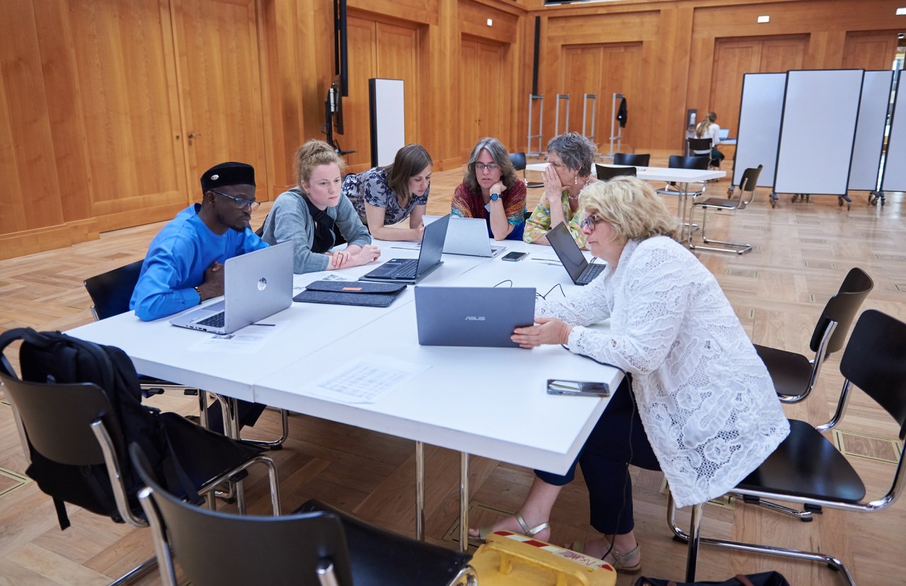 Sechs Teilnehmende sitzen im Weltsaal zusammen und arbeiten gemeinsam an Laptops.