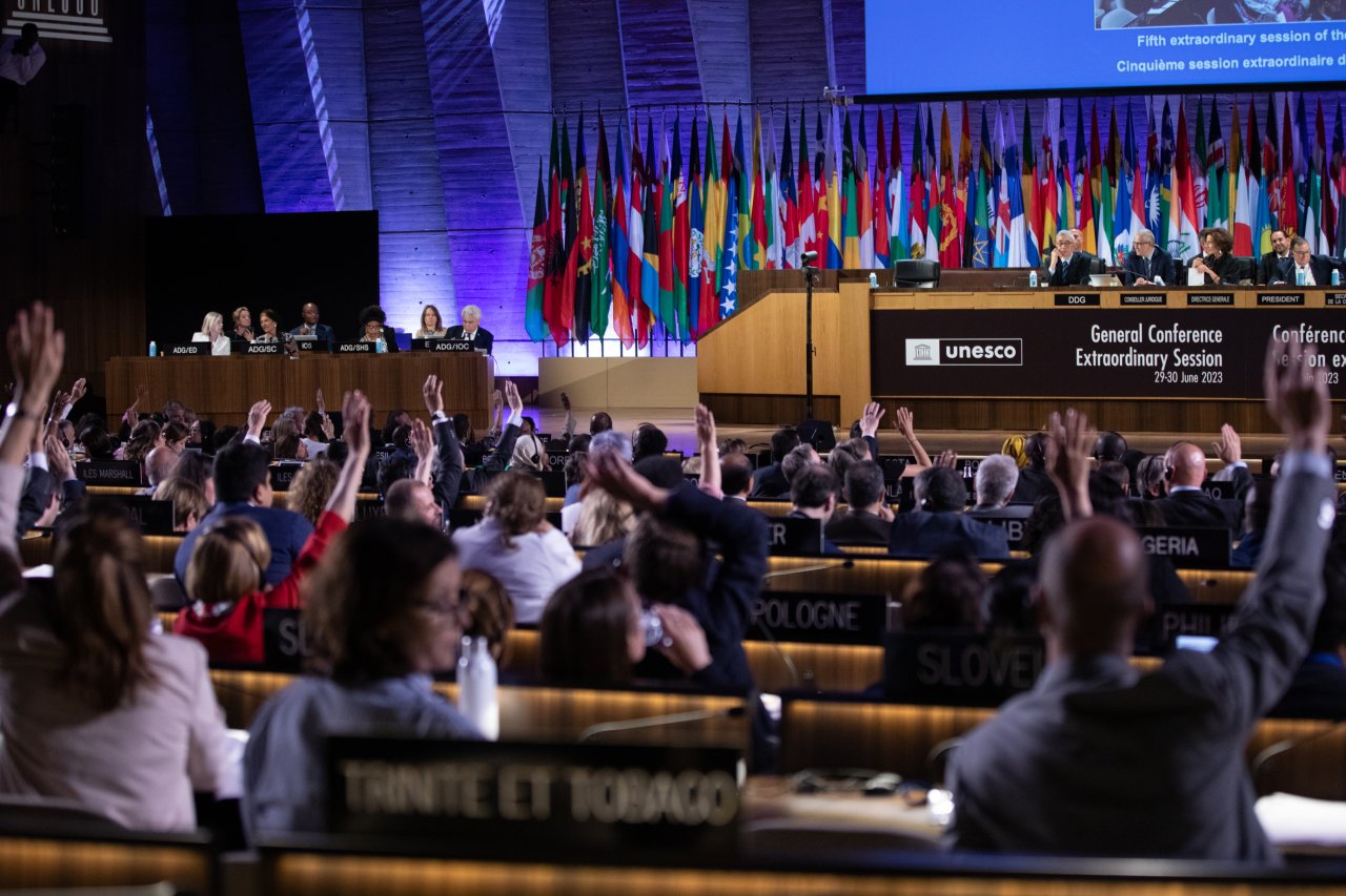 Zahlreiche Personen im Plenum der UNESCO-Generalkonferenz heben während einer Abstimmung ihre Hände.