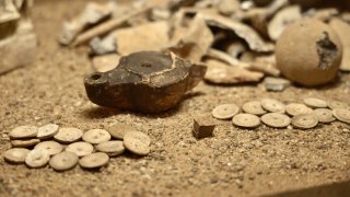 Auf sandigem Untergrund liegen antike Grabbeilagen, darunter kleine Spielsteine in Form von Münzen und ein Würfel.