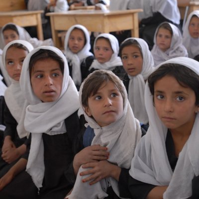 Das Bild zeigt etwa 15 Mädchen in einer Schule. Sie sitzen entweder auf Teppichen oder an Tischen. Sie alle tragen Hijab.